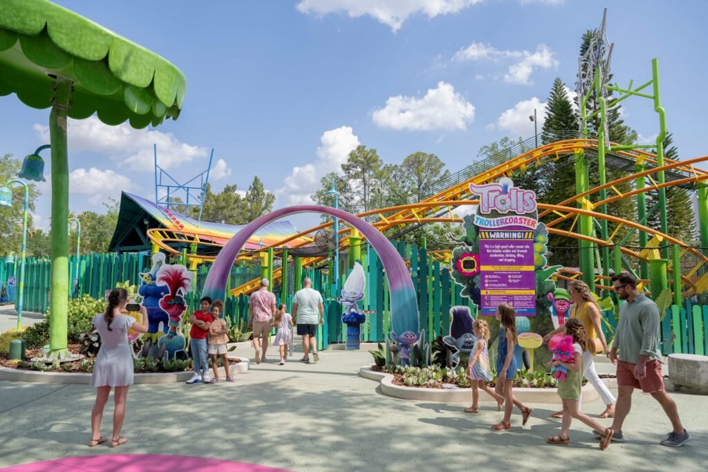 A vibrant and colorful rollercoaster at Universal Studios, with a sign reading “Trolls” in front of the entrance, surrounded by lush greenery and visitors enjoying the park.