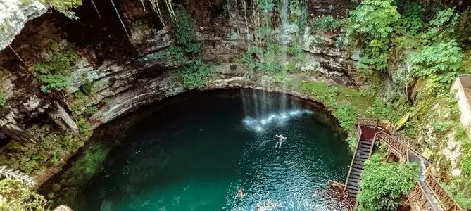 Aerial view of a natural cenote with crystal-clear water surrounded by lush greenery and a rocky overhang, with visitors swimming and a staircase leading down to the water.