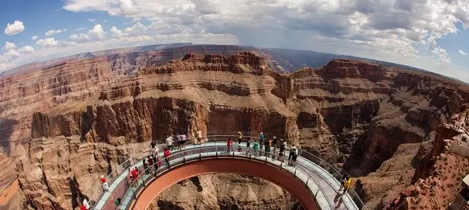 Visitors on a curved glass bridge extending over the Grand Canyon under a vast blue sky with clouds.