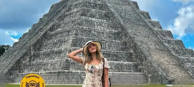 Traveler in floral dress standing before El Castillo pyramid at Chichen Itza, hand on forehead shading eyes, under a partly cloudy sky.