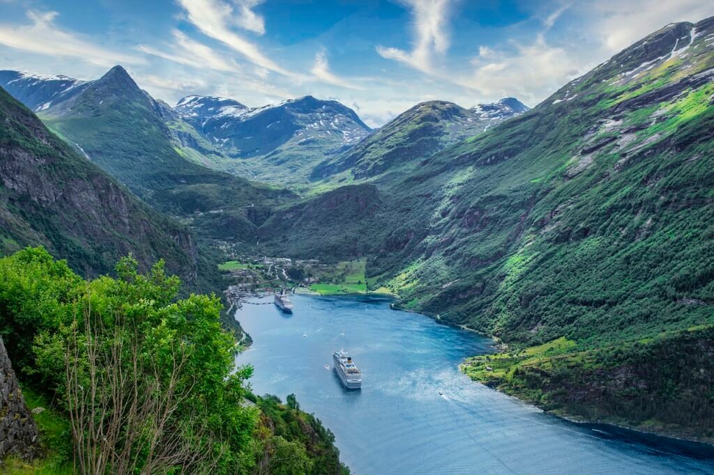 A panoramic view of a majestic fjord with steep green mountains and a small village by the shore, featuring a large cruise ship sailing through the vibrant blue waters, maybe your next surprise travel adventure!