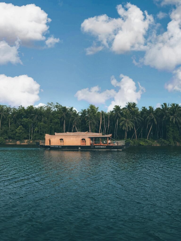 A traditional wooden houseboat floating on the calm waters of a river in Kerala, India, with lush green palm trees in the background under a clear blue sky. Soulful and compassionate travel destinations.