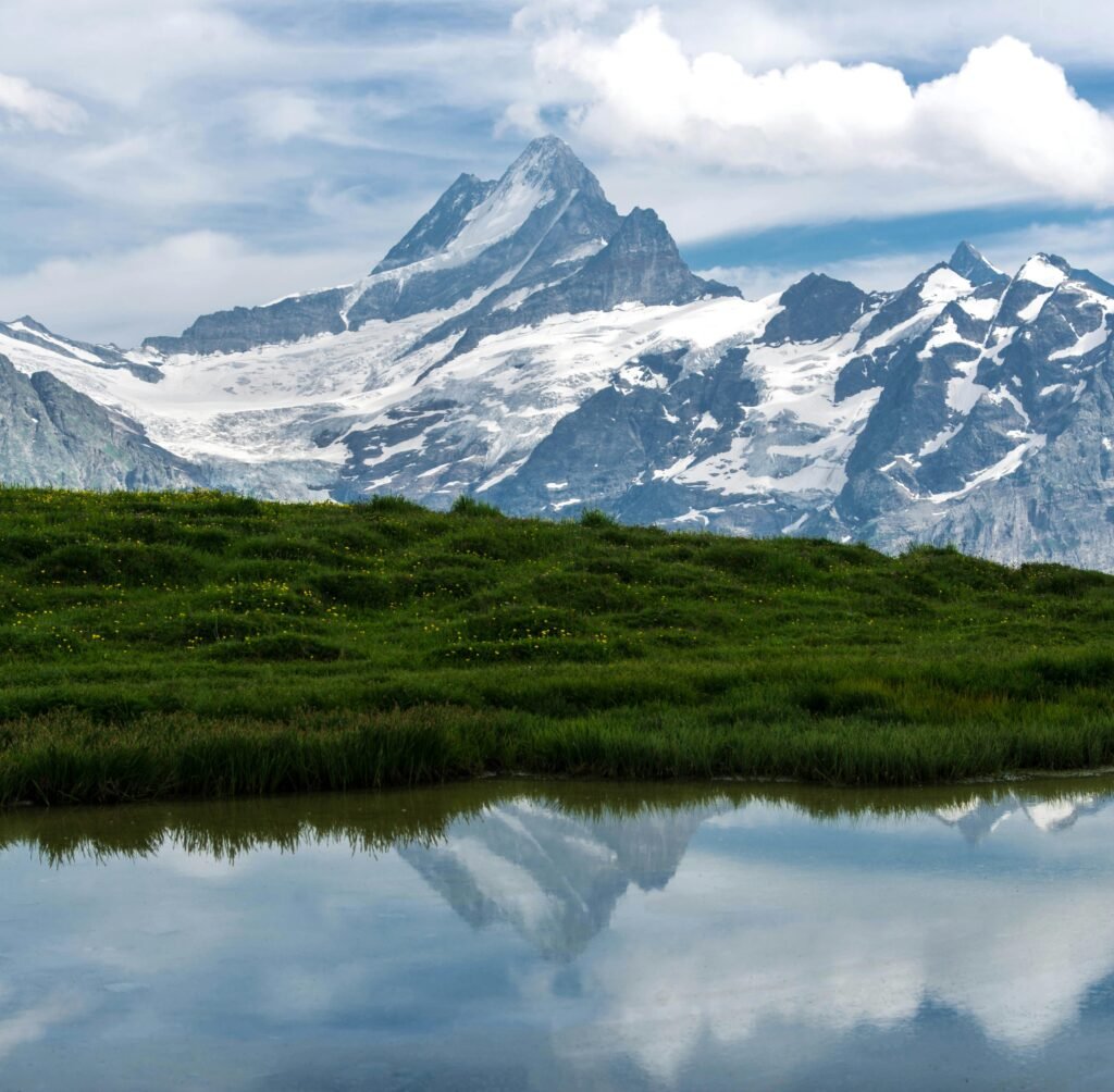 A tranquil Swiss landscape with a snow-capped mountain reflected in a still water body amidst green meadows under a partly cloudy sky.