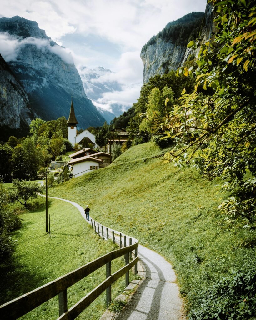 A person walking on a path through green fields towards a small village at the base of mist-covered mountains in Switzerland.