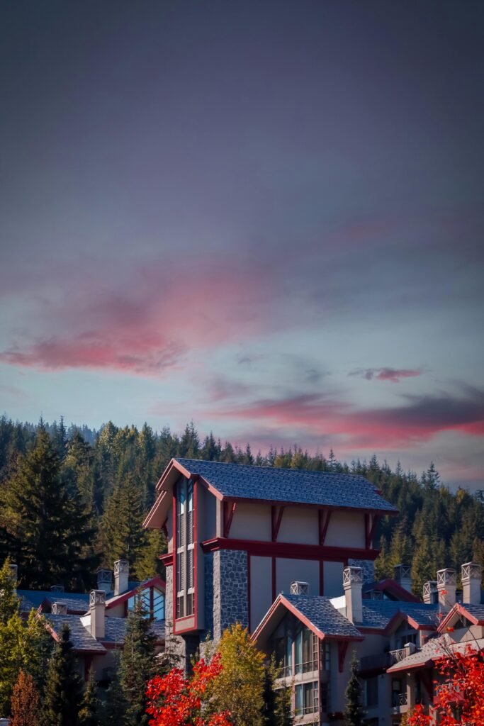 A multi-story house with a blue roof and red accents set against evergreen trees under a sky with pink and orange hues.