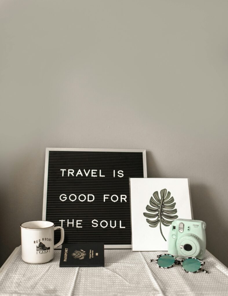 A still life composition on a white table featuring a letter board with the quote ‘TRAVEL IS GOOD FOR THE SOUL,’ next to a mint green instant camera, a candle labeled ‘BEACH,’ a passport, and two leaf-shaped earrings on top of an open book.