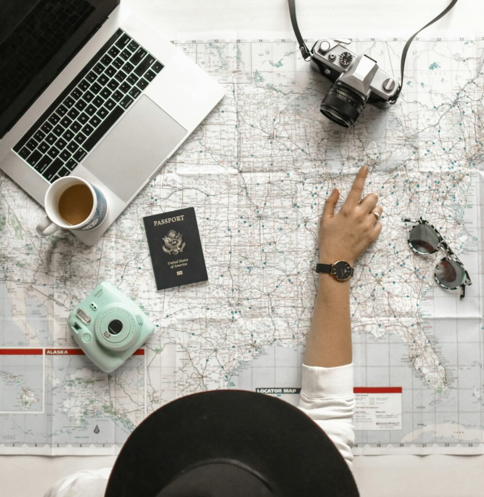 A person’s hand pointing at a location on a world map, surrounded by travel essentials including a laptop, camera, passport, coffee cup, and sunglasses.