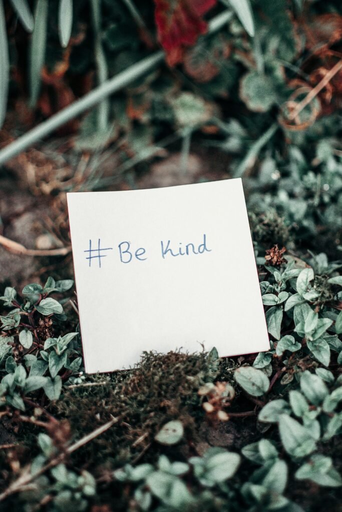 A white note with the message ‘#Be Kind’ written in blue ink, placed on a bed of green leaves and moss.