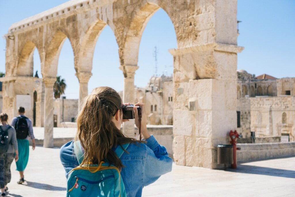 A person with long hair and a blue backpack taking a photo of ancient stone arches under a clear sky.