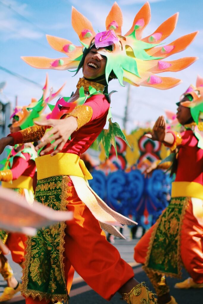 A group of performers in vibrant, colorful costumes with large flower-like headpieces dancing in a street parade.