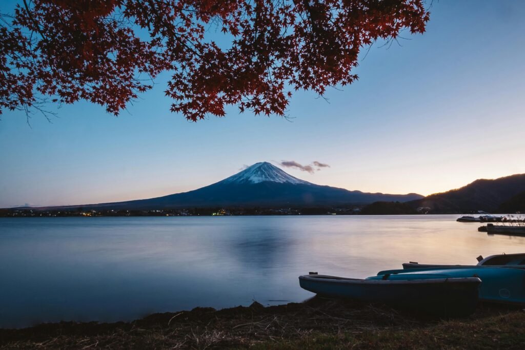 Twilight view of snow-capped Mount Fuji reflected in a calm lake with red maple leaves in the foreground and blue boats on the shore.