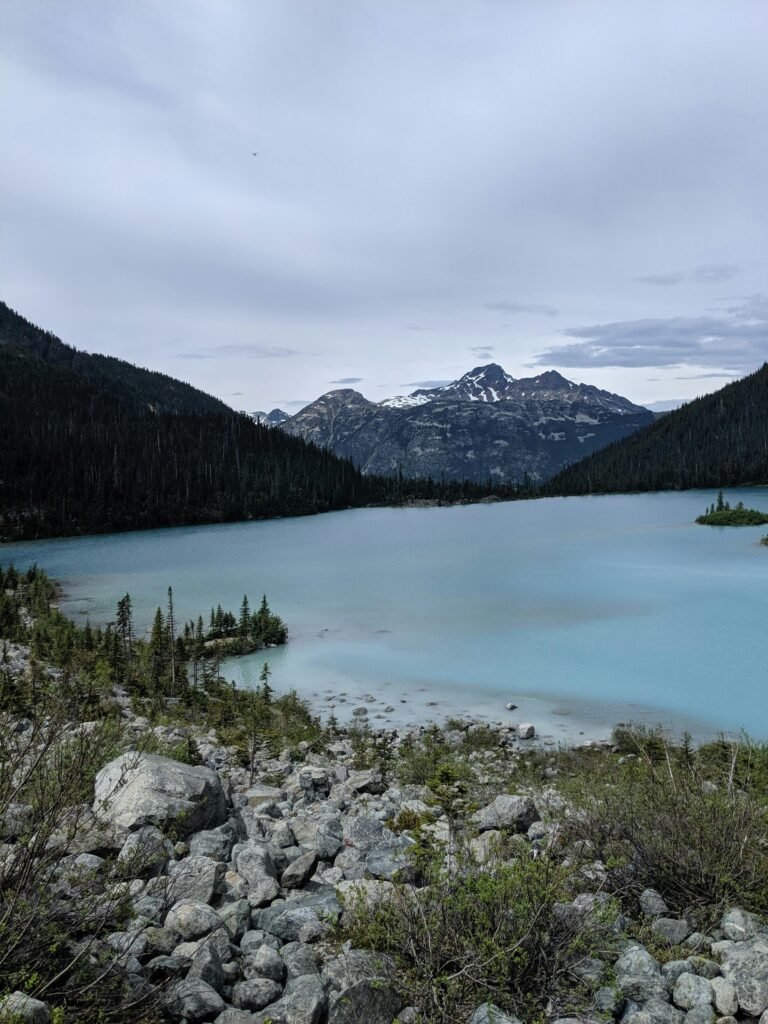 A tranquil turquoise lake nestled among lush green forests with majestic mountains looming in the background under a soft cloudy sky.