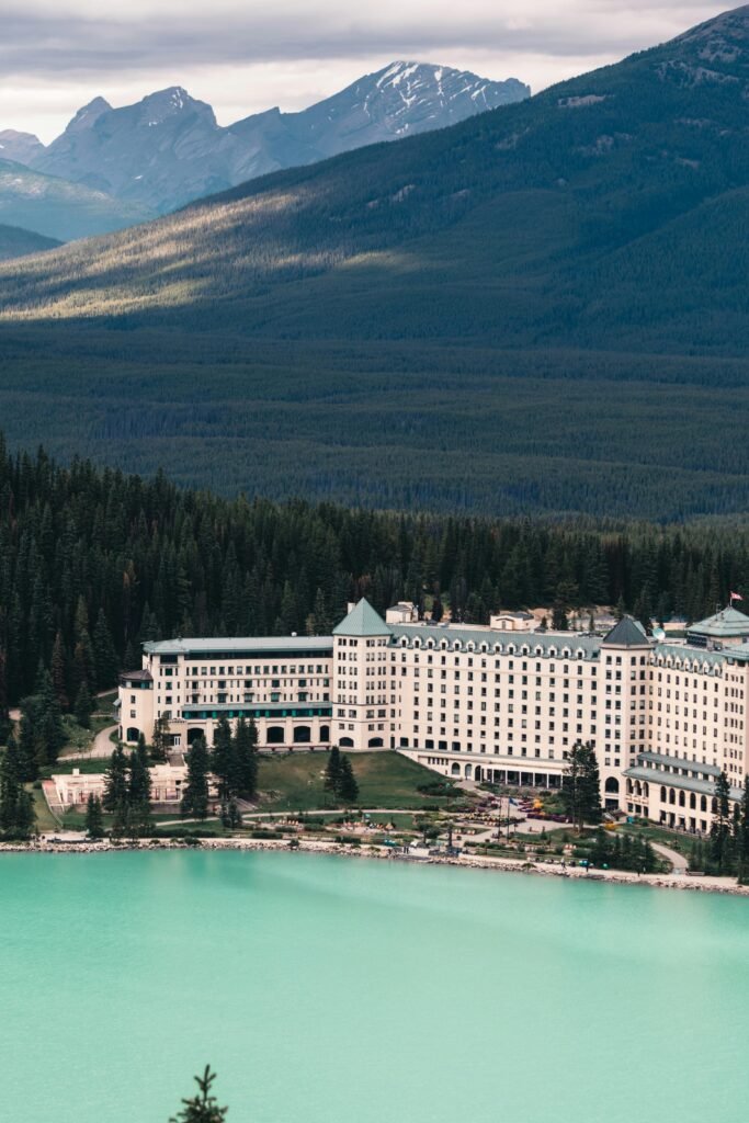 A large white hotel overlooking a turquoise lake set against forested mountains under a cloudy sky.