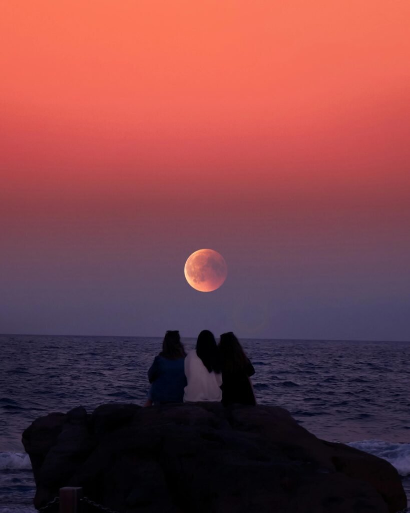 Three people sitting on a rock at the beach, silhouetted against a vibrant sunset with a full moon rising above the horizon, embodying travel with heart.