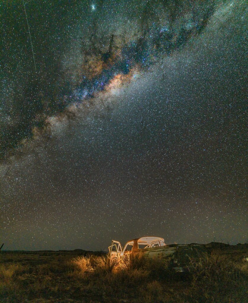 A starry night sky with the Milky Way galaxy visible above a grassy field, where a car with open doors is parked. Perfect for witnessing upcoming celestial events.