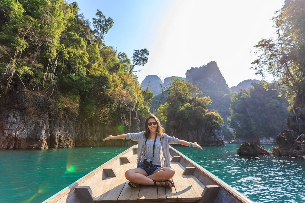 A person sitting cross-legged at the front of a wooden boat, with outstretched arms, facing a body of water surrounded by tall rock formations and lush greenery.