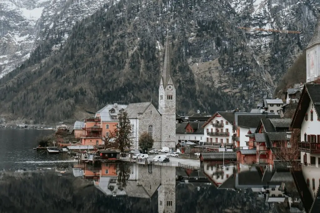 A serene lakeside village with a prominent church spire, nestled against a backdrop of steep mountains, reflecting perfectly on the calm water surface. A perfect destination for a surprise travel adventure!