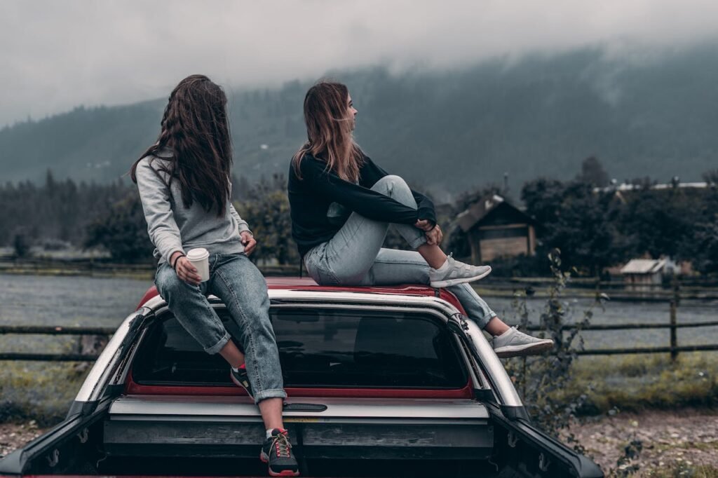 Two people sitting on the hood of a red car facing away from the camera, looking towards misty mountains in a rural setting.