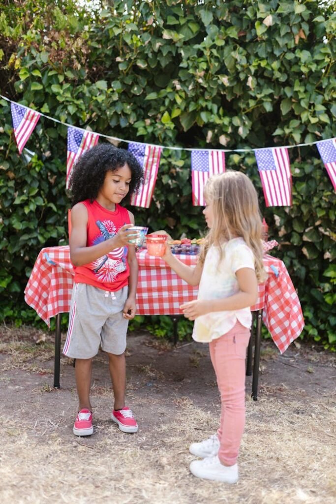 Two children at an outdoor Labor Day celebration, standing by a table with a red and white checkered tablecloth, serving themselves food.