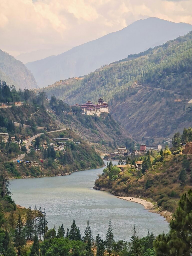 A scenic view of a river winding through a mountainous landscape in Bhutan, with a prominent monastery perched on the hillside. Soulful and compassionate travel destinations.