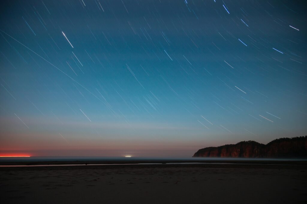 A serene night sky with star trails over a tranquil beach and forested cliffs, hinting at the Earth’s rotation. Perfect for witnessing upcoming celestial events.