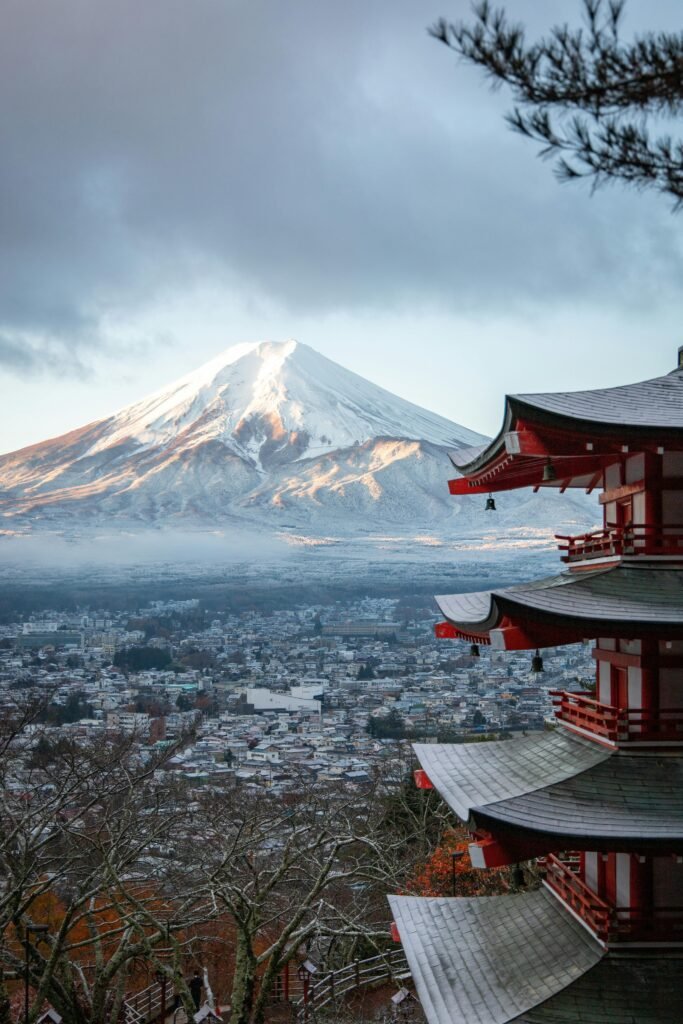 Snow-capped Mount Fuji illuminated by sunlight behind a shadowed multi-tiered pagoda, with a misty town below.