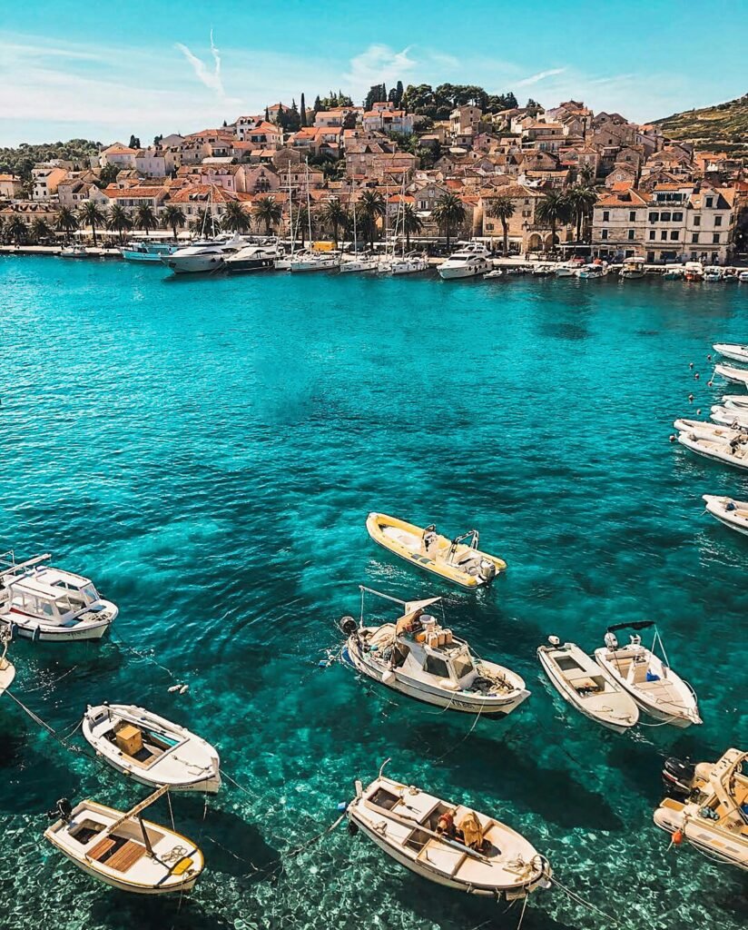 A vibrant coastal scene with multiple boats floating on clear turquoise waters, with a picturesque hillside town in the background under a clear blue sky.