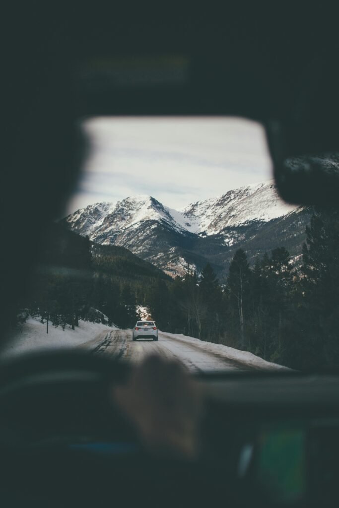 View from inside a car showing another car on a snow-covered road leading towards distant mountains.