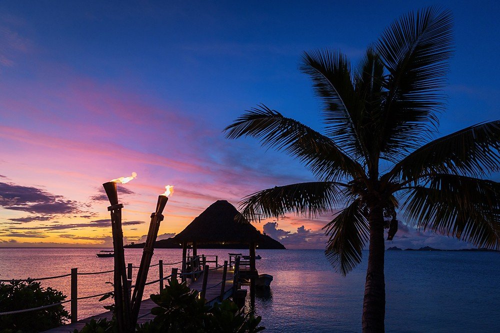 A serene twilight scene at Likuliku Lagoon with a vibrant sky transitioning from pink to deep blue, featuring silhouettes of palm trees, a traditional torch, and a distant island.