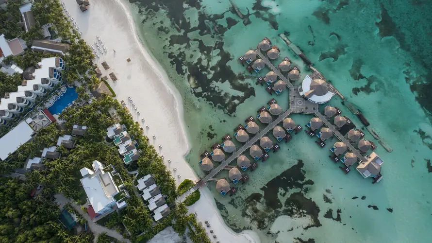 Aerial view of a luxury resort in Playa del Carmen with overwater bungalows extending into the turquoise sea, surrounded by lush greenery and a white sandy beach.