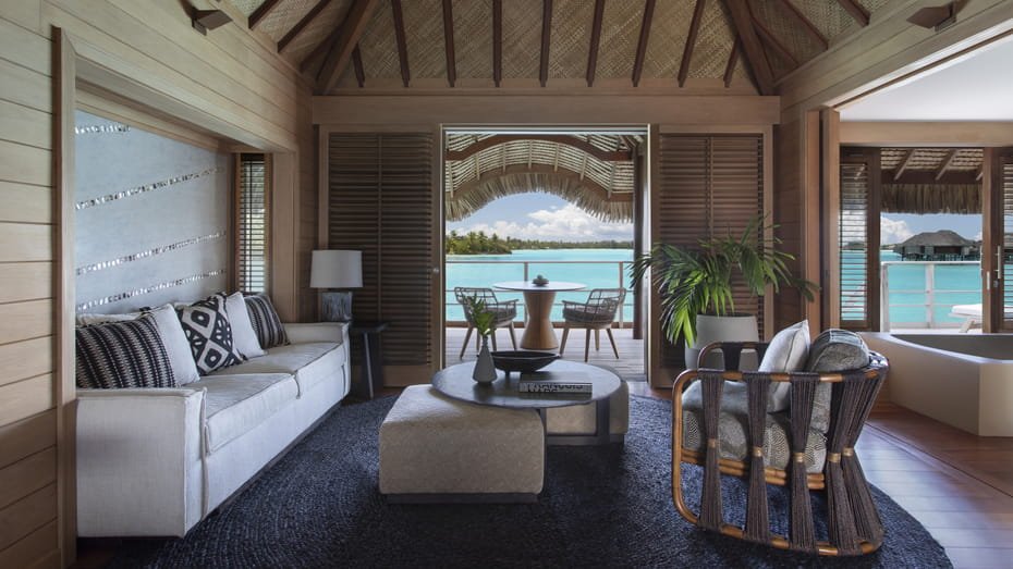 Interior view of a luxurious overwater bungalow with elegant furnishings and large windows showcasing a panoramic view of the turquoise lagoon and Mount Otemanu.