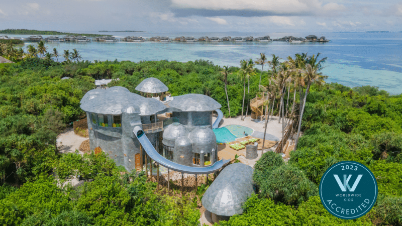 Aerial view of The Den at Soneva Jani, featuring a large, whimsical building with a silver dome-like roof surrounded by lush greenery, with a long blue waterslide extending towards an outdoor pool, set against the backdrop of the clear turquoise waters of the Maldives.