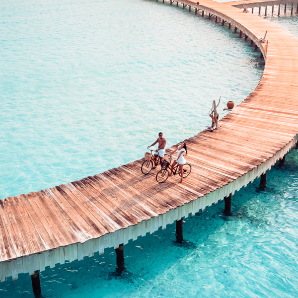 A couple riding bicycles on a curved wooden pier over clear turquoise waters with tropical marine life visible beneath.