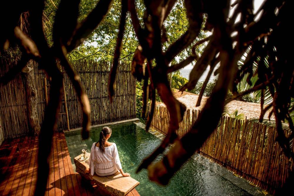 A person sits at the edge of a serene private pool surrounded by a bamboo fence and lush greenery, with sunlight filtering through the foliage.