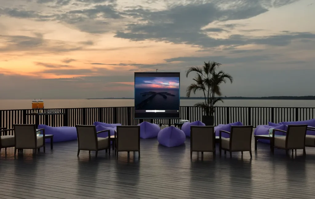 A serene outdoor lounge area at dusk with a large screen displaying a sunset, overlooking a calm sea.