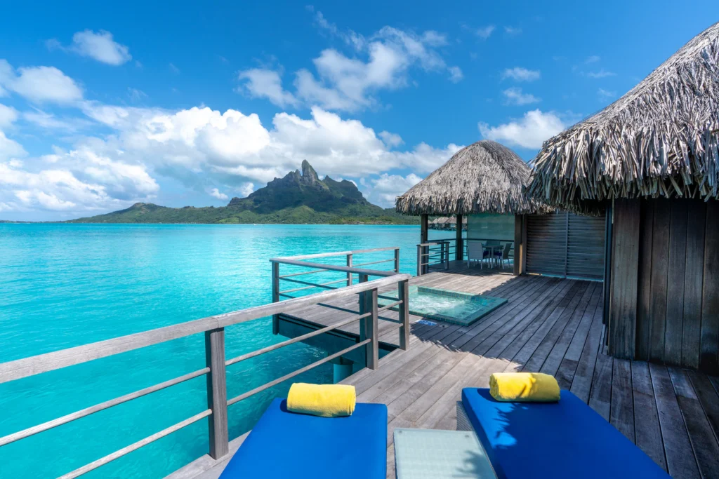 Overwater bungalows with thatched roofs extend over the clear turquoise waters of a lagoon, with a view of Mount Otemanu in the distance under a blue sky with scattered clouds.