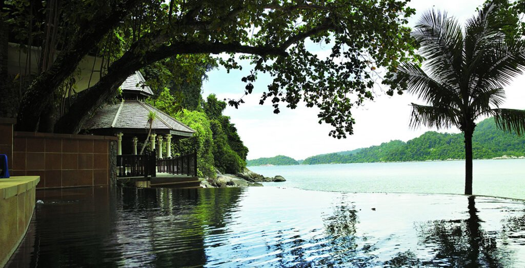 An infinity pool blending with the sea at Pangkor Laut Resort, flanked by lush greenery and traditional wooden structures.