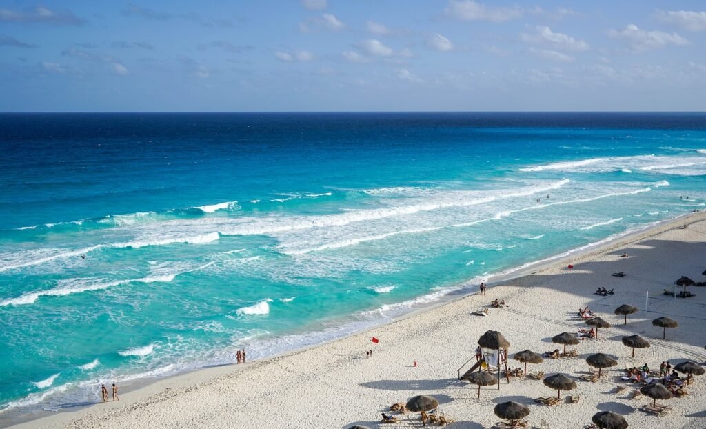 A panoramic view of a beach in Cancún with turquoise waters and white sands, dotted with umbrellas and beachgoers under a clear blue sky.