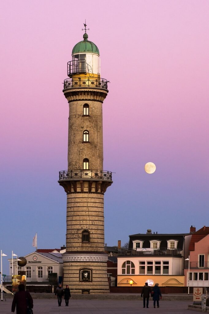 A tall, cylindrical lighthouse with a green dome stands against a twilight sky with shades of pink and purple, with a full moon visible in the background. Harvest Supermoon Eclipse