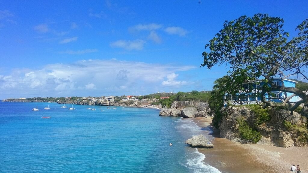 A panoramic view of Curaçao’s coastline with clear blue skies, turquoise waters, and a rocky shoreline. A gazebo perched on the cliffside overlooks several boats in the water, and people are visible on the small beach below. Caribbean and West Indies Vacation