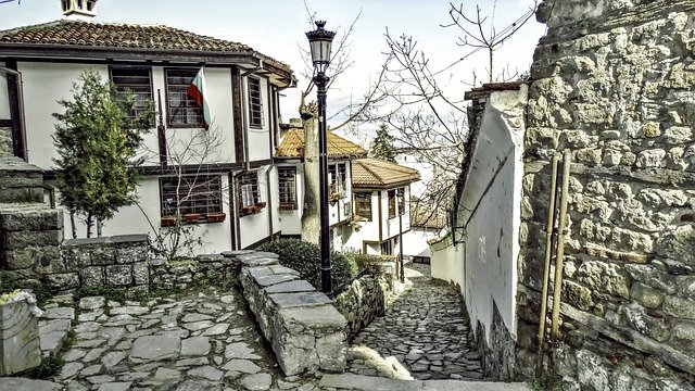 A narrow cobblestone street in Plovdiv, flanked by traditional Bulgarian houses with white walls and wooden frames, a stone wall to the right, and a vintage street lamp.