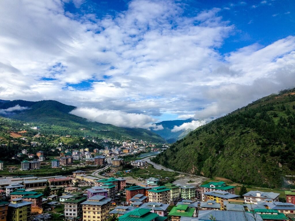 A panoramic view of a valley in Bhutan with a mix of traditional and modern buildings nestled among green, forested hills under a blue sky with scattered clouds.