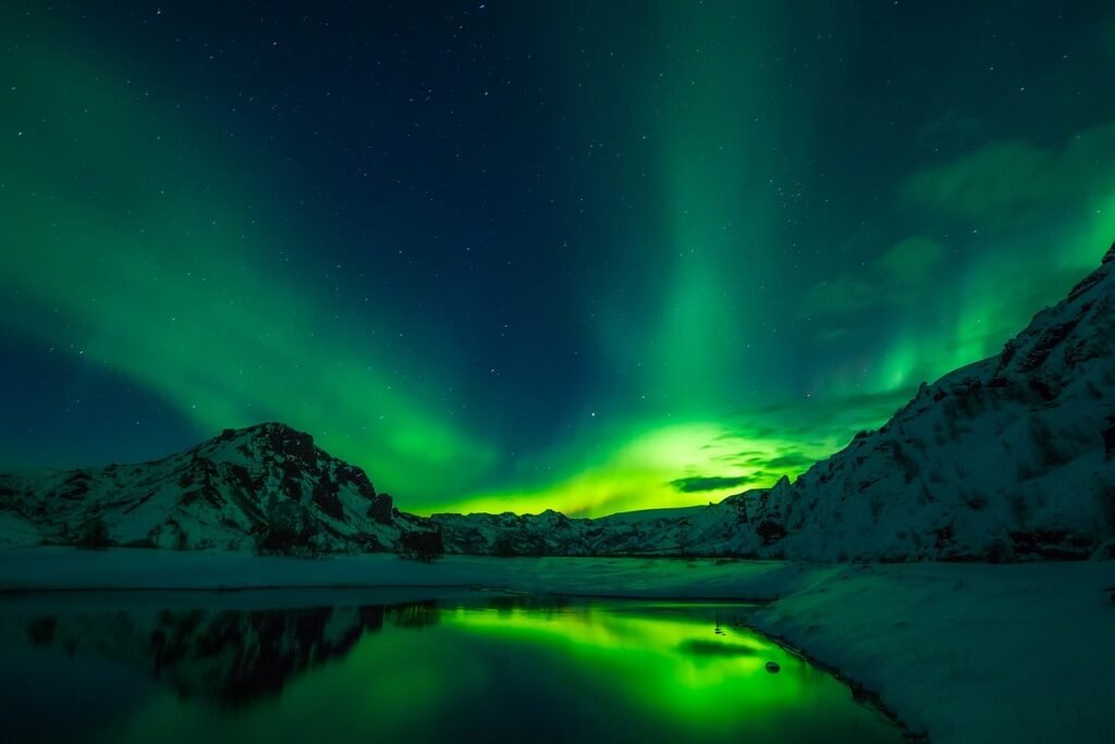 Aurora Borealis with vibrant green hues over a snow-covered landscape and still water in Iceland.