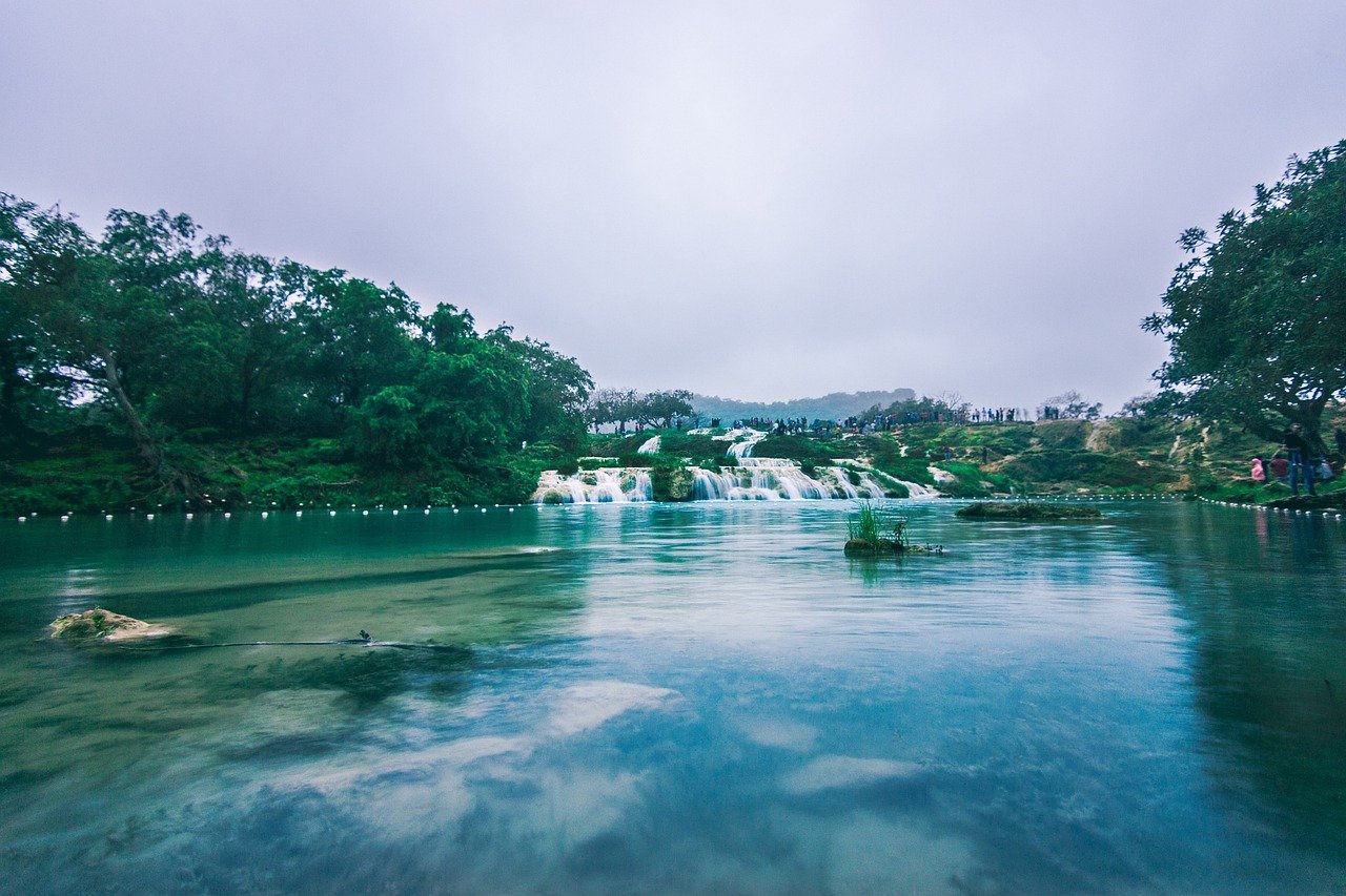 Unique travel destination Salalah with waterfalls and lush greenery. A tranquil scene with multiple small waterfalls cascading into a clear blue lake surrounded by lush greenery under an overcast sky.