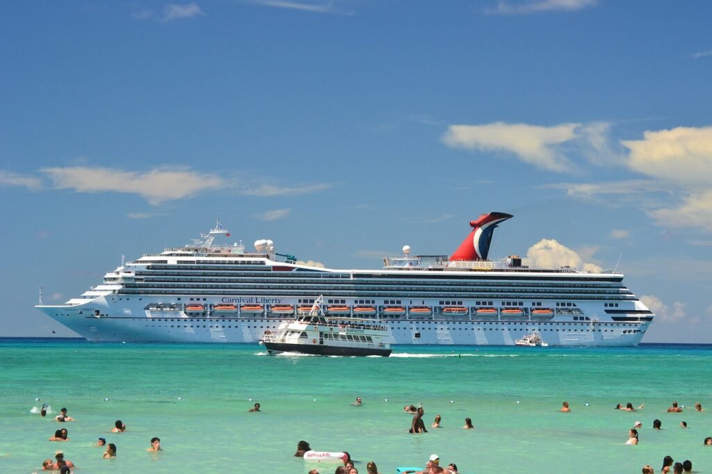 Carnival Liberty cruise ship anchored near a beach with people swimming in clear blue waters under a sunny sky.