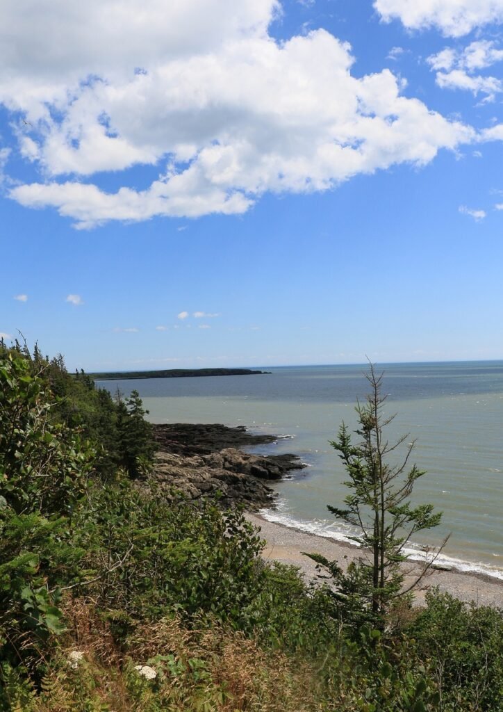  A scenic coastal landscape with a clear blue sky dotted with clouds, overlooking a calm sea. The coastline features rocky outcrops and lush greenery.