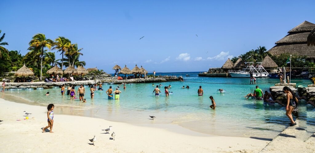 Vibrant beach scene with people enjoying the water and sand, thatched-roof structures, and palm trees in Mexico.