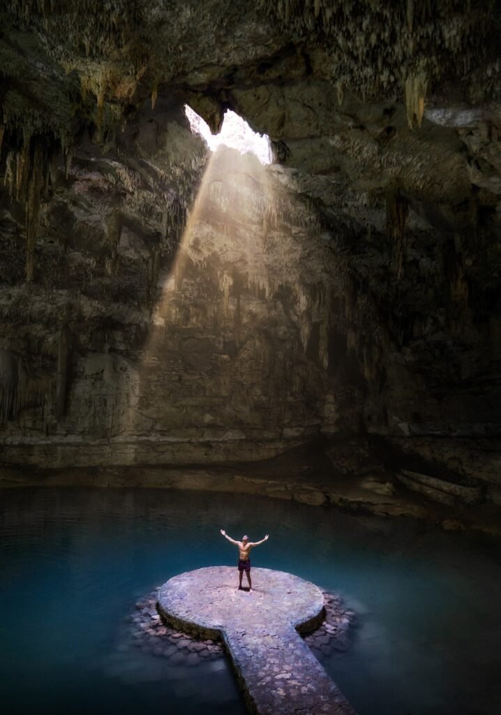 mexico, cave, cenote. A person stands with arms outstretched on a circular platform in the center of an underground water-filled cavern, illuminated by a shaft of light from an opening above.