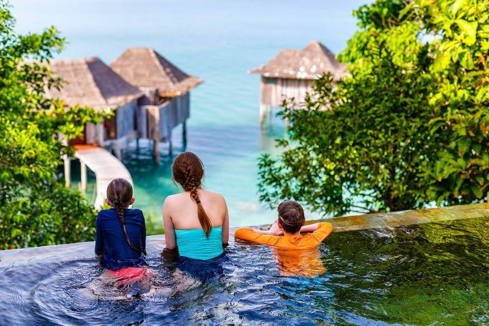 Three individuals seated at the edge of an infinity pool overlooking a seascape with overwater bungalows.