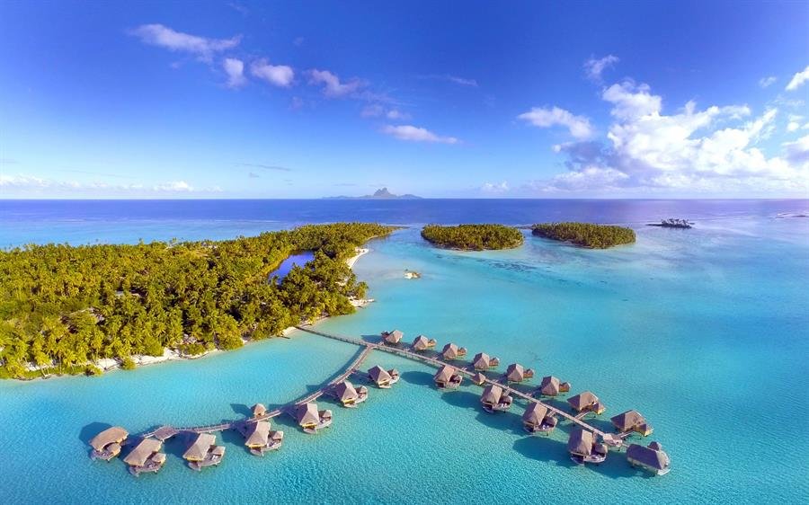 Aerial view of Le Taha’a by Pearl Resort showcasing overwater bungalows extending into a turquoise lagoon, surrounded by lush greenery and a distant mountainous backdrop.